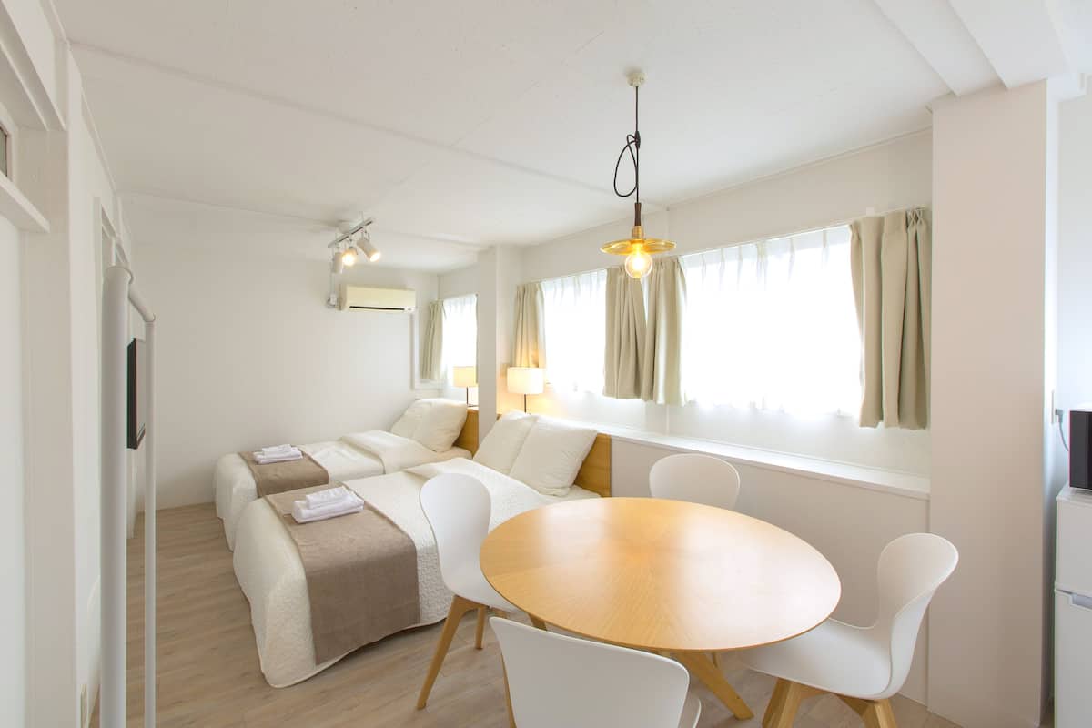 A modern, well-lit room with a round dining table, two white chairs, a sofa with a beige throw, and a window with curtains.
