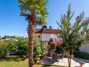 A courtyard with a palm tree, potted plants, a patio with a chair, and a building with a tiled roof.