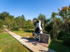 Un barbecue extérieur en pierre, doté d’une cheminée, entouré d’herbe et d’arbres.