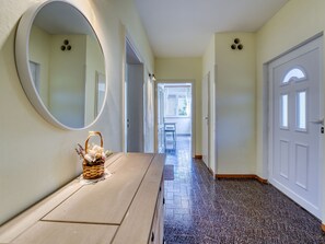 Un pasillo con un banco de madera, un espejo redondo y una cesta con flores. Hay puertas a ambos lados y una ventana al final.