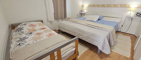 A bedroom with two beds, a wooden floor, a window with curtains, and a wall-mounted lamp.