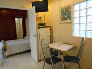 A compact room with a bed, a small kitchenette, a dining table with chairs, a TV mounted above the kitchenette, and a window with a view.