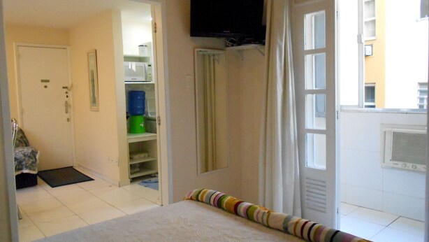 Une chambre à coucher comprenant un lit, un téléviseur fixé au mur, une armoire et une fenêtre donnant sur des immeubles.