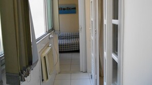 Un couloir étroit, avec un lit à motifs en damier visible au fond, un radiateur sur la gauche et une porte entrouverte sur la droite.