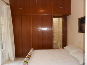 A bedroom with a large wooden wardrobe, a bed with white bedding, and a bathroom visible through an open door.