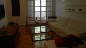 A living room with a sofa, a glass coffee table, a TV, and a window with blinds. - Gohouse - Atlantica 702 B (Rio de Janeiro)