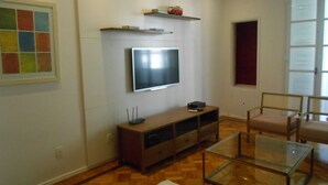 A living room with a flat-screen TV mounted on the wall, a wooden entertainment unit below, a glass coffee table, and two wicker chairs. - Gohouse - Atlantica 702 B (Rio de Janeiro)