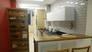 A compact kitchen with a small island, two wooden chairs, and a bookshelf filled with various items. - Gohouse - Atlantica 702 B (Rio de Janeiro)