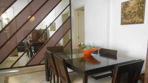 A dining area with a glass table, wooden chairs, and a plant centerpiece. A staircase is visible in the background. - Gohouse - Anibal 604 (Rio de Janeiro)