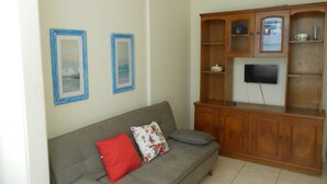 Una sala de estar con un sofá gris, dos cuadros enmarcados, un mueble de madera con un televisor y piso de baldosas.
