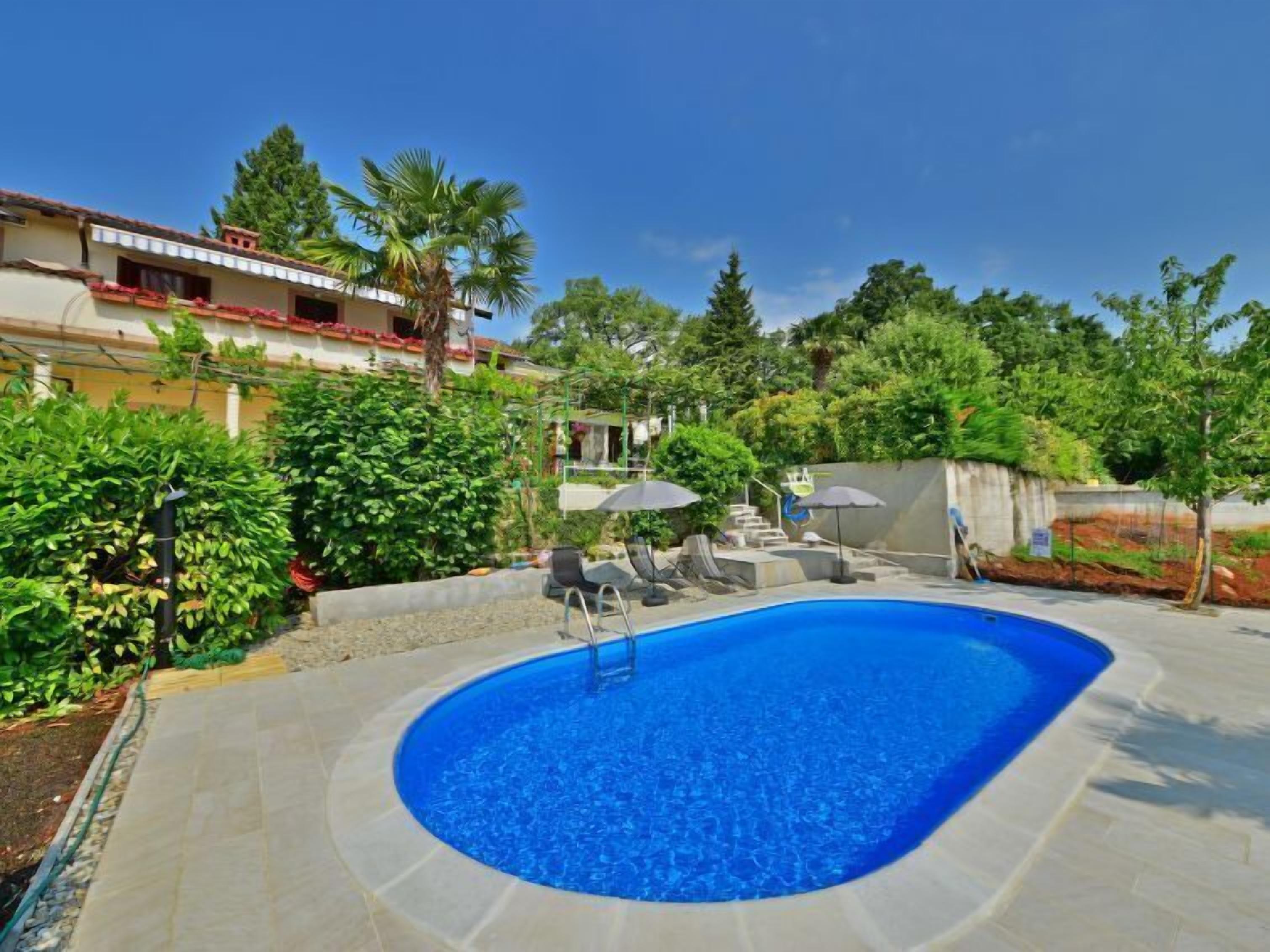 Piscina com deck ladrilhado, rodeada por vegetação e um edifício com telhado vermelho.