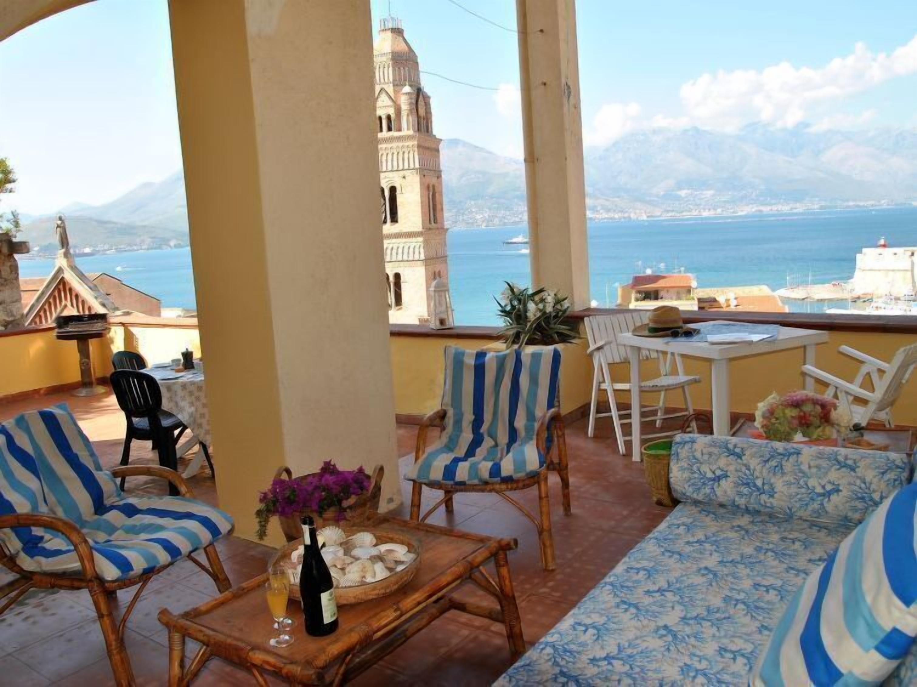 A terrace with wicker furniture, a table with a bottle and glasses, and a view of the sea and mountains.