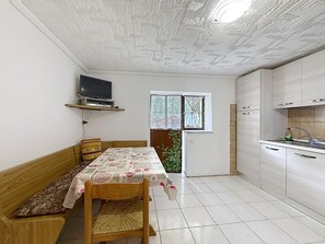 A compact kitchen with a dining area, a television mounted on a shelf, and a window with a view of greenery.