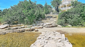Un hotel costero con un sendero de piedra que lleva al mar, rodeado de exuberante vegetación y un cielo azul despejado.