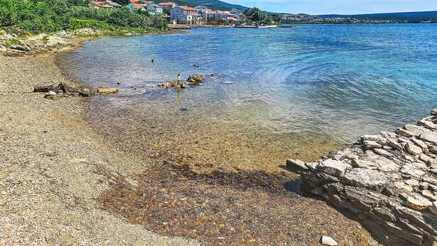 Paisaje costero con agua cristalina, una costa rocosa y edificios al fondo.