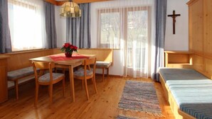 Una stanza con pavimento in legno, un tavolo da pranzo con sedie, una panca, un letto, una croce al muro e ampie finestre con tende.