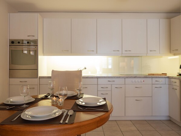 Een moderne keuken met witte kasten, een ingebouwde oven, en een eethoek voor twee personen met een houten tafel, borden, glazen en bestek.