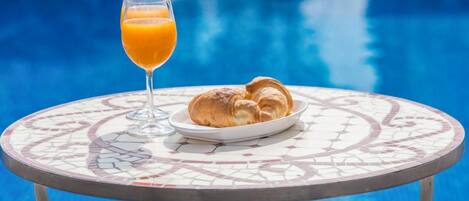 A table with a glass of orange juice and croissants by a poolside.