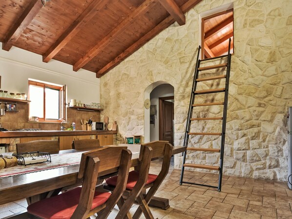 Una cocina rústica con paredes de piedra, vigas de madera, una zona de comedor con sillas tapizadas en rojo y una escalera apoyada contra la pared.