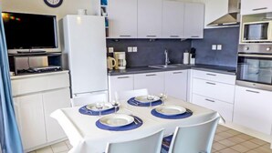 Une cuisine compacte avec des meubles blancs, un four à micro-ondes, une machine à café et une table à manger pour deux personnes, agrémentée de sets de table bleus.