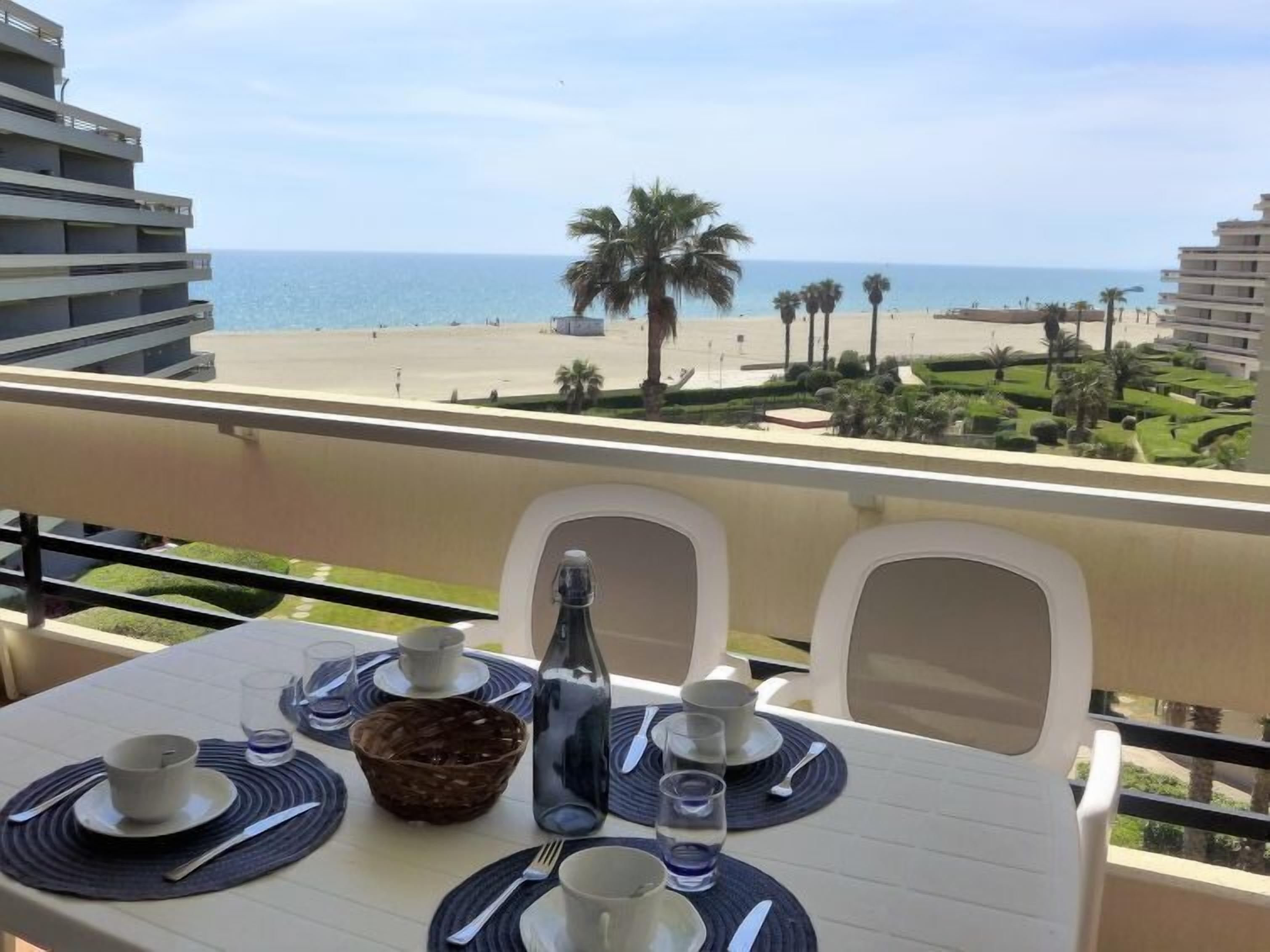 A balcony table set with dinnerware, a bottle, and a basket, overlooking a beach with palm trees and a clear sky.