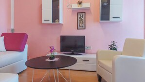 Olohuone, jossa on vaaleanpunainen seinä, valkoiset huonekalut, litteänäyttöinen televisio ja pieni pöytä, jonka päällä on koriste-esine.