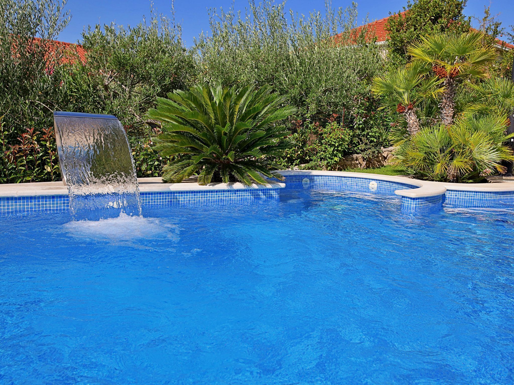 A swimming pool with a waterfall feature, surrounded by lush greenery and a tiled edge.