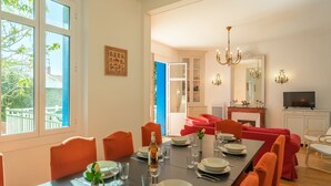 Een eetruimte met een lange tafel gedekt voor vier personen, omringd door oranje stoelen. Daarboven hangt een kroonluchter en er is een televisie in de achtergrond zichtbaar.