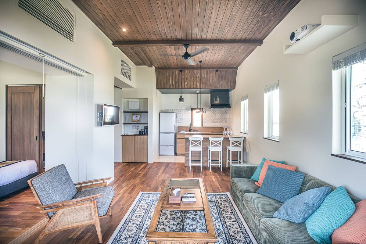 Uma sala de estar moderna com teto de madeira, área de cozinha com banquetas e uma grande janela que oferece luz natural.