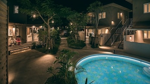 Vista nocturna de una zona residencial con piscina, senderos y casas con ventanas iluminadas.