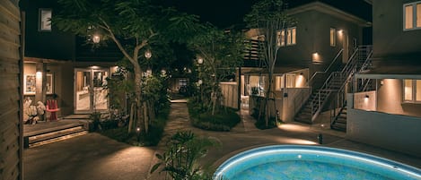 Vista nocturna de una zona residencial con piscina, senderos y casas con ventanas iluminadas.