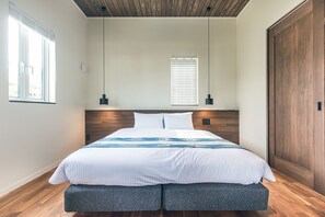 A modern bedroom with a wooden headboard, a large bed with a patterned bedspread, two bedside lamps, and a large window with blinds. - Mihana Onna-Son (Onna)