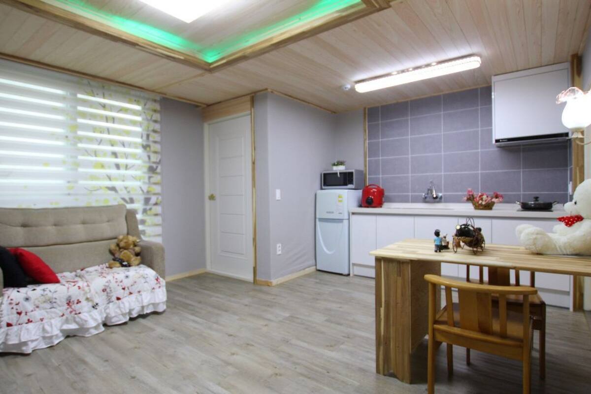 A cozy living room with a sofa, a small table, and a teddy bear. There is a kitchen area with a microwave, a kettle, and a sink.
