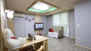 Une pièce chaleureuse avec un plafond en bois, une télévision, un canapé beige agrémenté d’un coussin rouge, une petite table sur laquelle est posé un ours en peluche, et une fenêtre avec des stores.