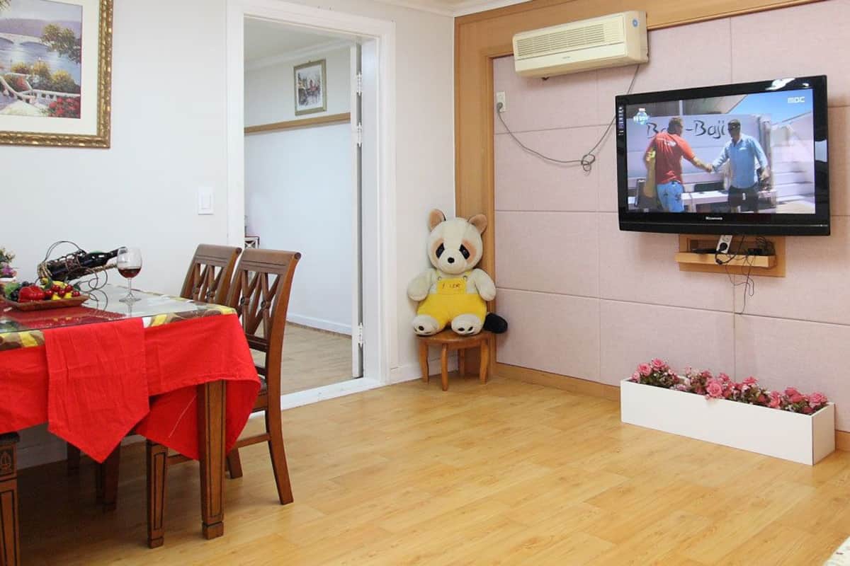 A living room with a dining table set for two, a television mounted on the wall, a sofa, and a decorative bear figure.