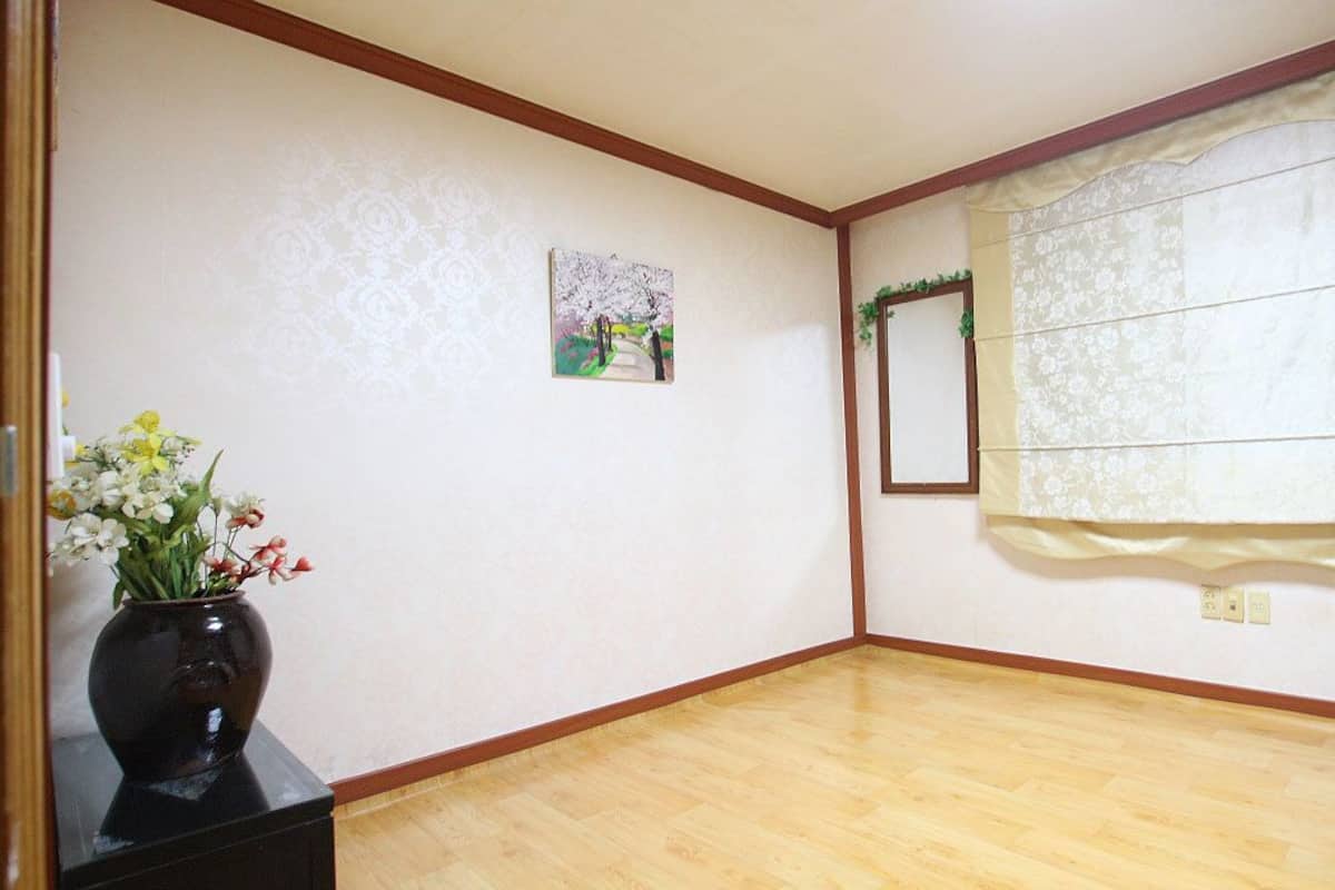 A room with a wooden floor, a vase with flowers, a framed picture, and a window with a curtain.