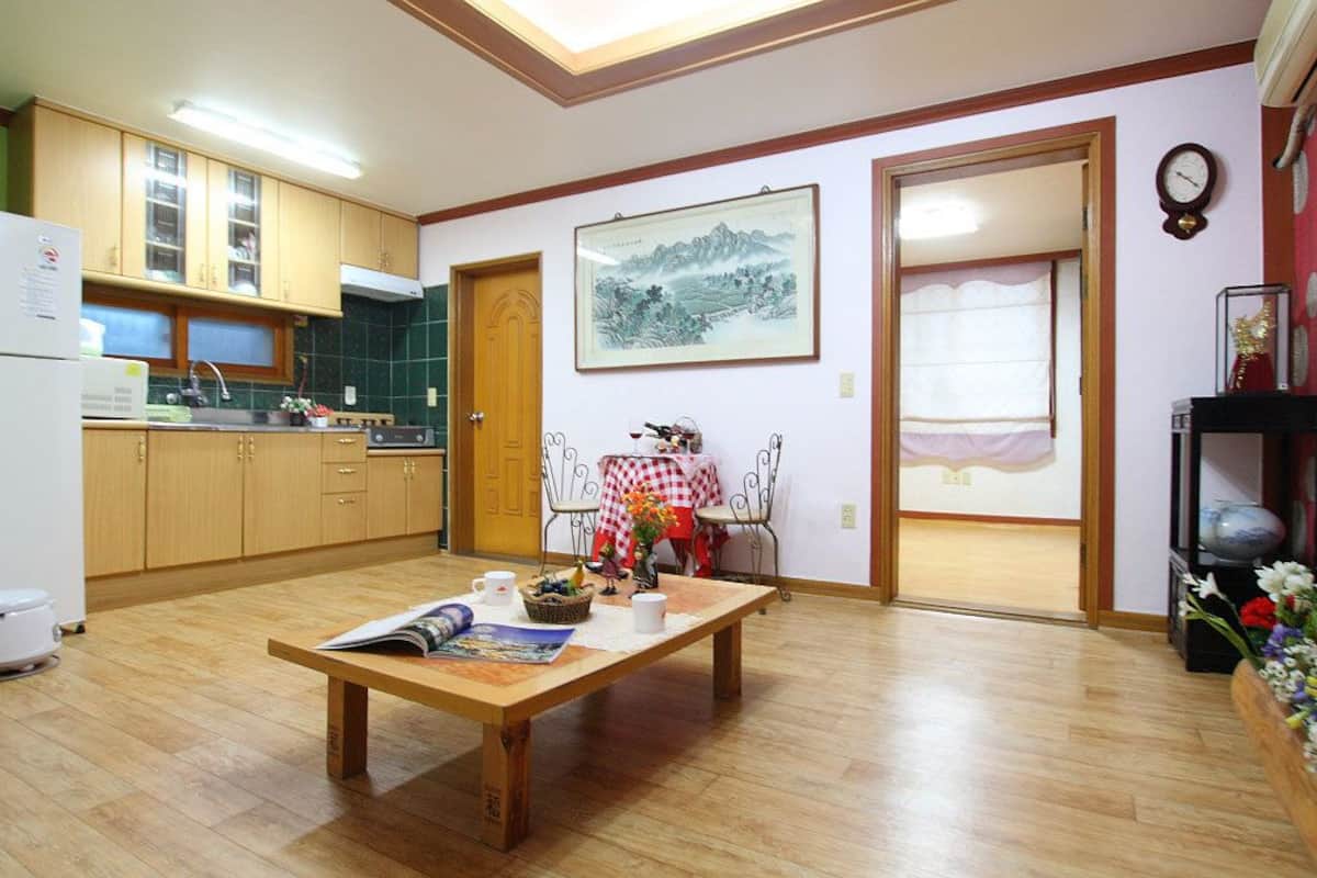 A compact kitchen with wooden cabinets, a dining table with a red and white checkered tablecloth, and a framed landscape painting on the wall.