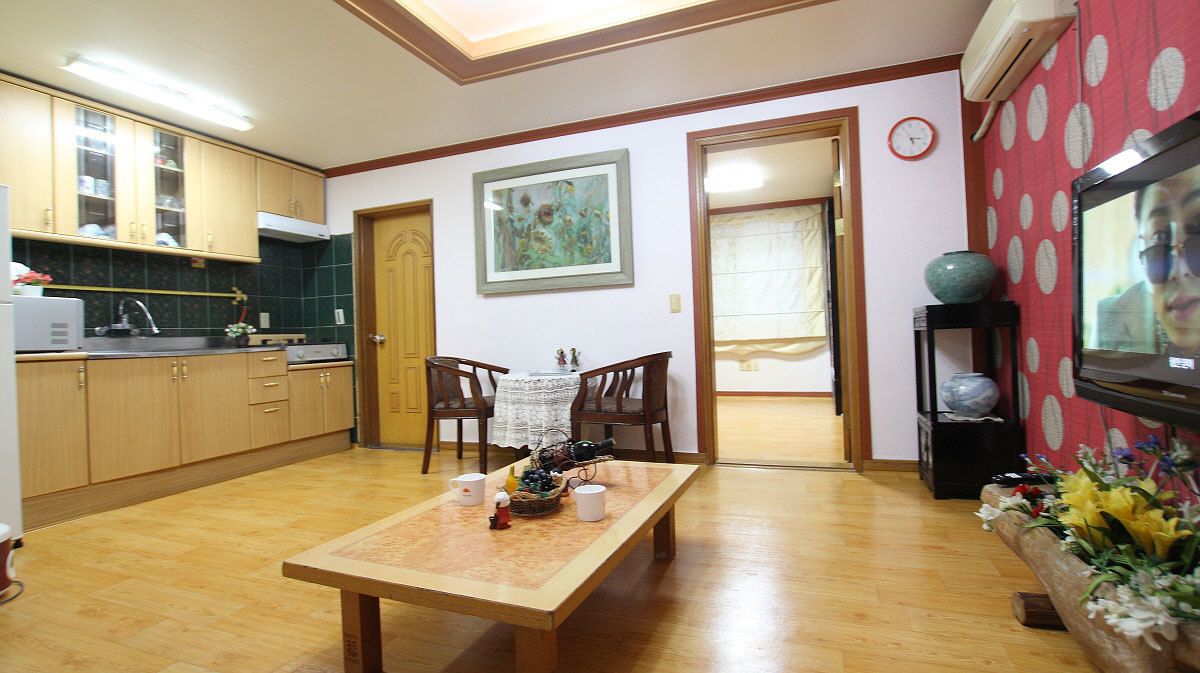 A kitchen with wooden cabinets, a dining table with chairs, a painting on the wall, and a television on the right.