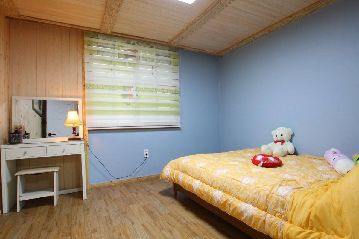A bedroom with a wooden desk, a bed with a yellow bedspread, a teddy bear, and a window with blinds.