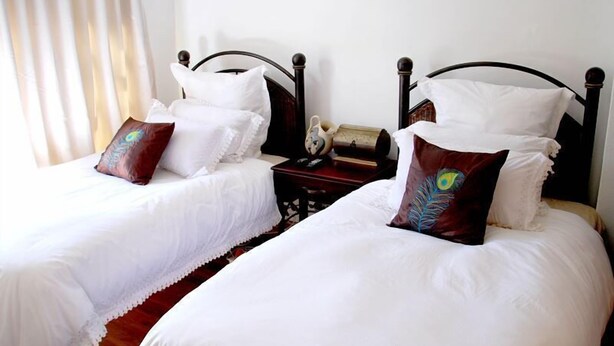 Two twin beds with white bedding and decorative pillows, a wooden nightstand with a lamp and a book, and a window with curtains. - Lapeng Habo Bantu (Mafeteng)