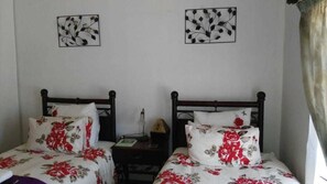 A room with two single beds, each with floral bedding and a wooden headboard. There are two bedside tables with lamps and a small decorative item on each. The walls are plain with two framed floral artworks. - Lapeng Habo Bantu (Mafeteng)
