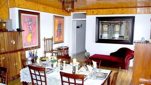 A dining room with a wooden table set for four, surrounded by chairs. There are framed artworks on the walls, a red sofa, and a mirror. - Lapeng Habo Bantu (Mafeteng)