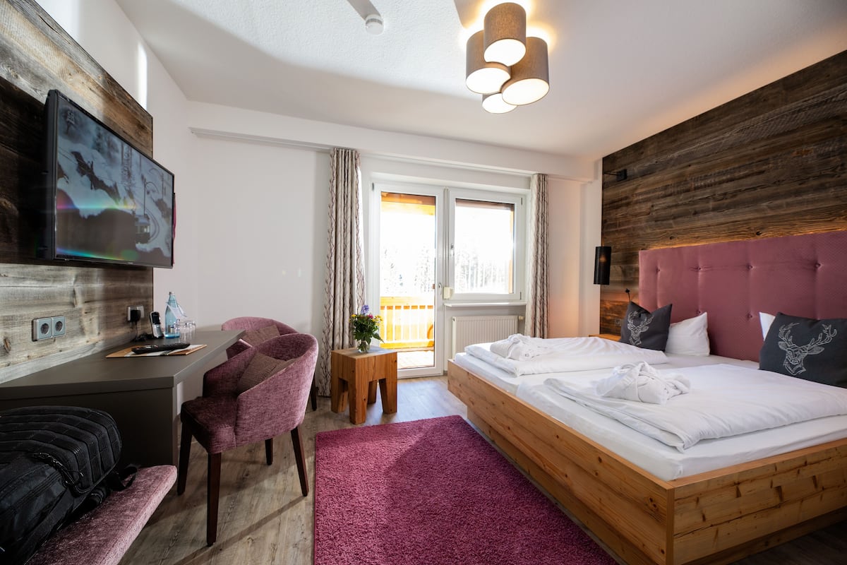 Een moderne slaapkamer met een houten hoofdbord, een bed met witte lakens, een bureau met een laptop, een stoel en een aan de muur bevestigde televisie.