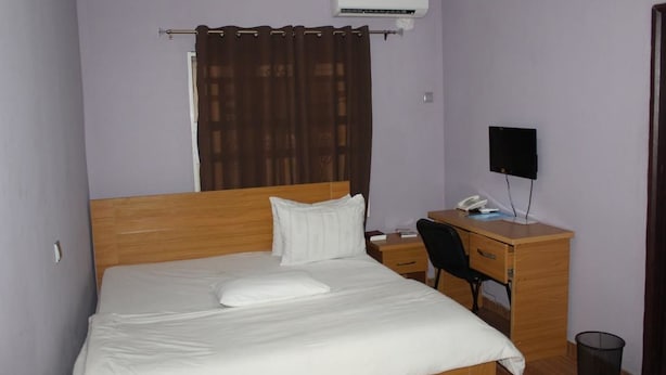 A neatly made bed with a white comforter and pillows, a wooden desk with a chair, a black speaker, and a small trash bin. - Rendezvous Hotel (Abuja)