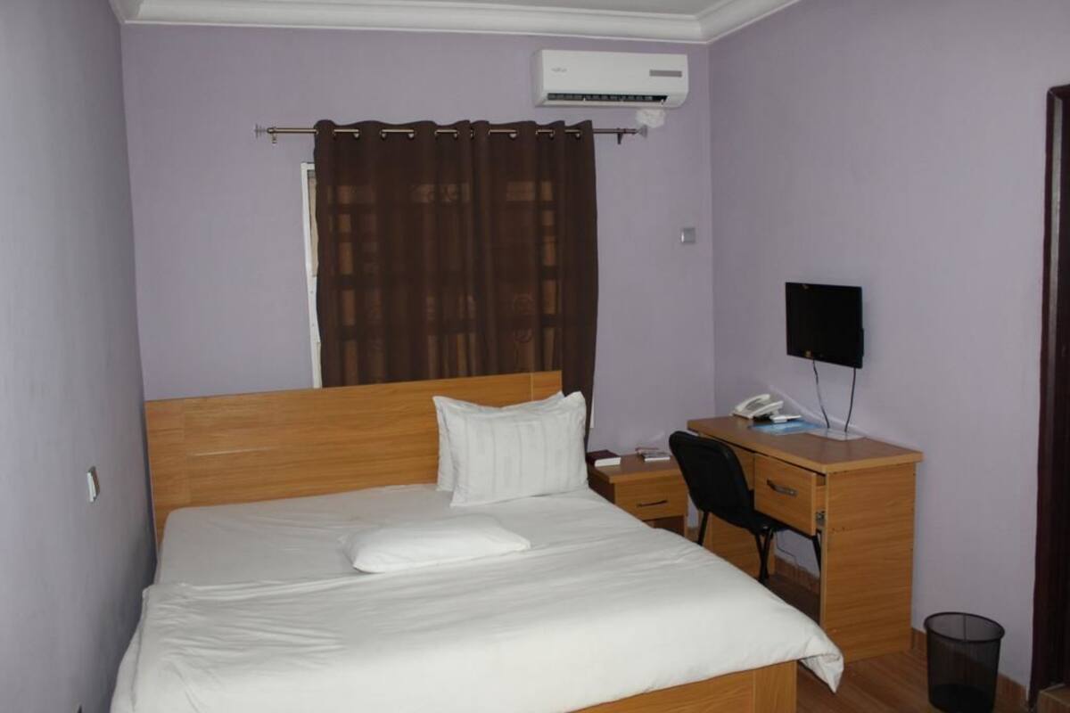 A neatly made bed with a white comforter and pillows, a wooden desk with a chair, a black speaker, and a small trash bin.