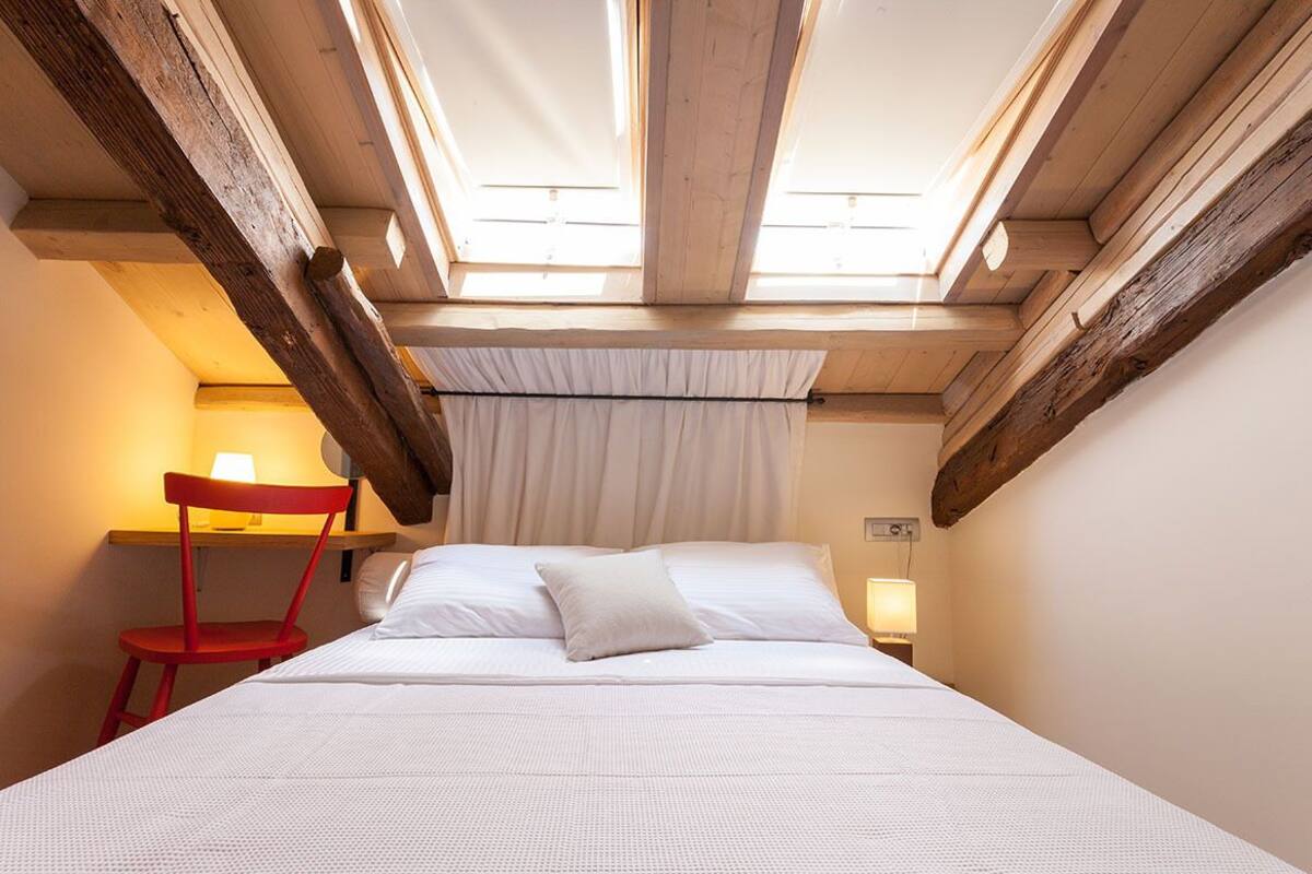 Ein Zimmer mit einem Bett, einem roten Sessel, einer Lampe und einem Fenster mit Holzbalken.