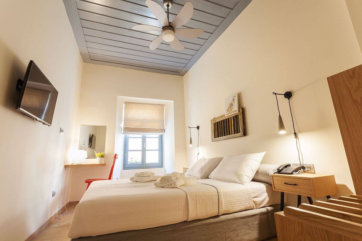 Ein Schlafzimmer mit einem Bett, einem Deckenventilator, einem Nachttisch mit Lampe, einem an der Wand befestigten Fernseher und einem Fenster mit Jalousien.