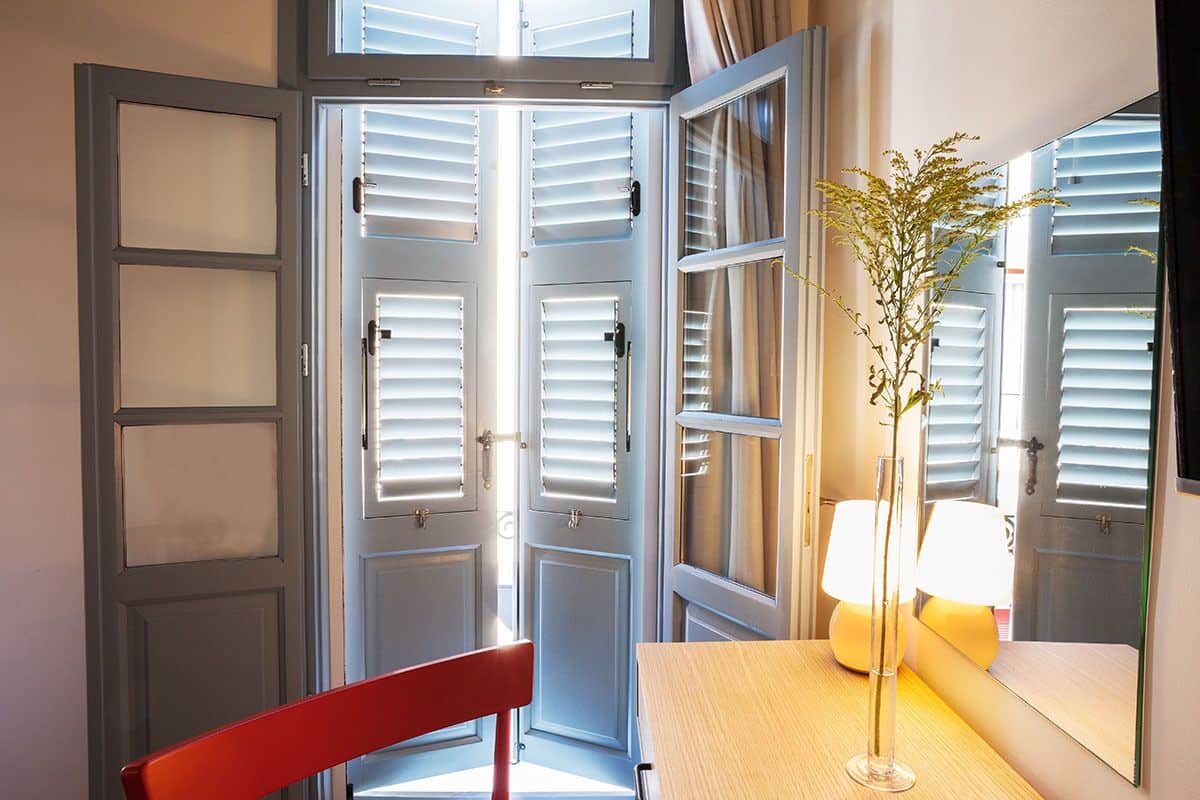 Ein Zimmer mit einem Holztisch, einem roten Stuhl, einer Vase mit Pflanze und einem Spiegel, der eine Tür mit Jalousien reflektiert.
