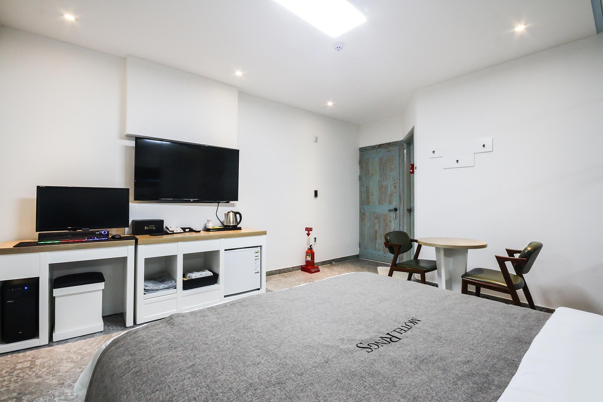 A modern hotel room with a flat-screen TV, a small kitchenette, a dining area with a table and chairs, and a bed with a grey blanket.