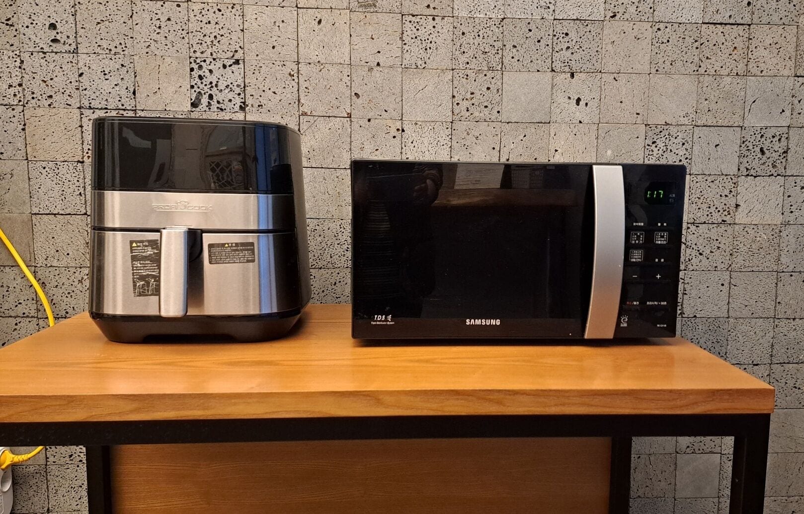 Una cafetera de acero inoxidable y un microondas negro sobre una mesa de madera, frente a una pared de azulejos con textura.